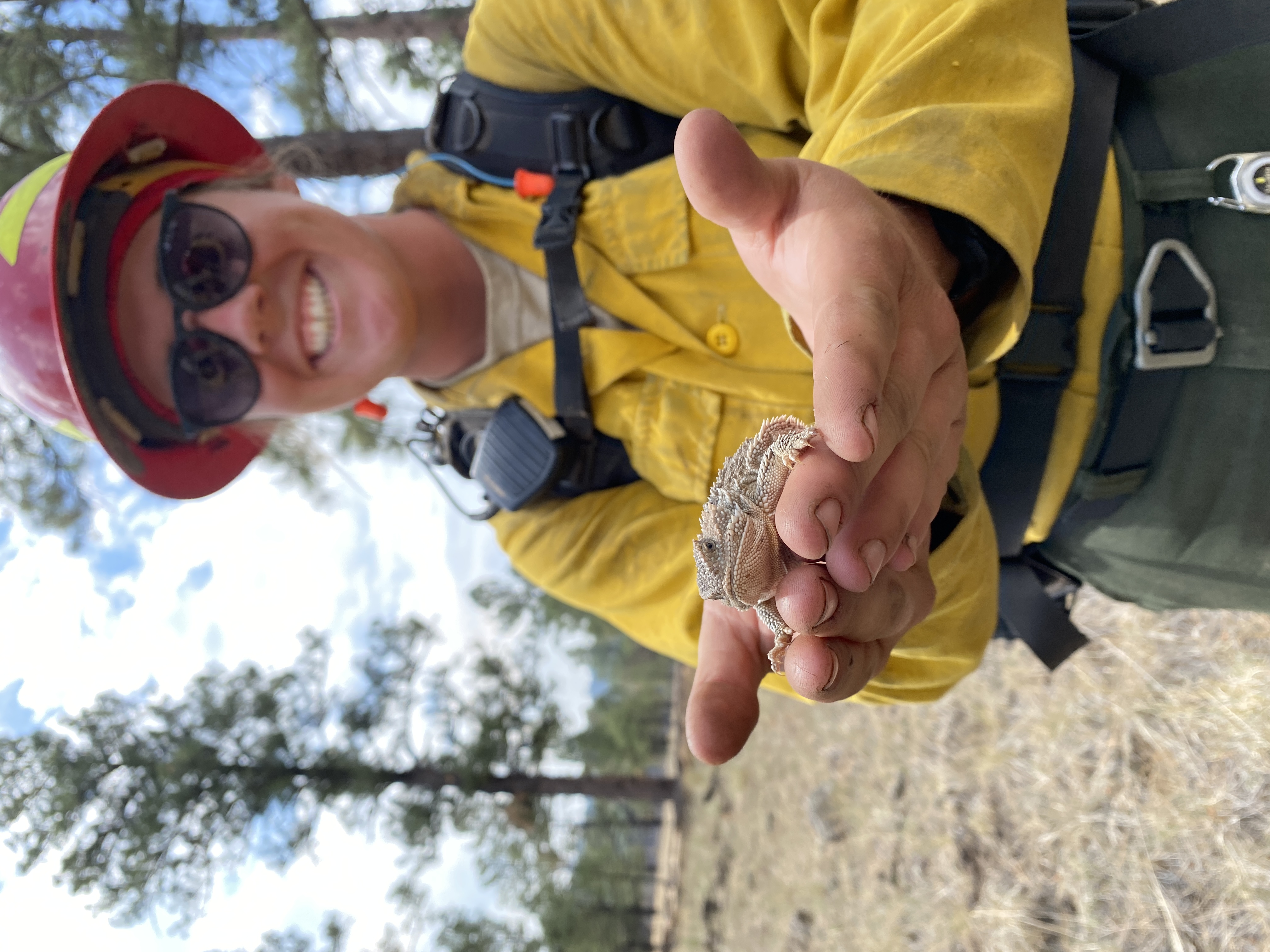 A woman in fire gear standing and holding a lizard out to the camera. 